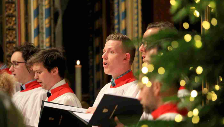 With a Christmas tree partially blocking the view on the right of the image, we see four singers of the Abbey Choir at Westminster Abbey in their white robes with red vestments, singig and holding hymn sheets