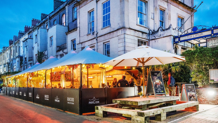 Outdoor seating area of The Clifton Pavilion, surrounded by black barriers, with large umbrellas and string lights. Wooden picnic tables are in the foreground, and the background features a historical building with large windows and white walls, under a twilight sky.