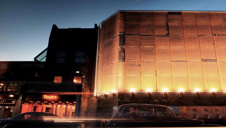 A city street at dusk, featuring a modern building with a glowing lattice facade illuminated in warm lights. To the left, a sign reading Theatre is seen in the entrance of an adjacent building. A classic car is parked along the street.