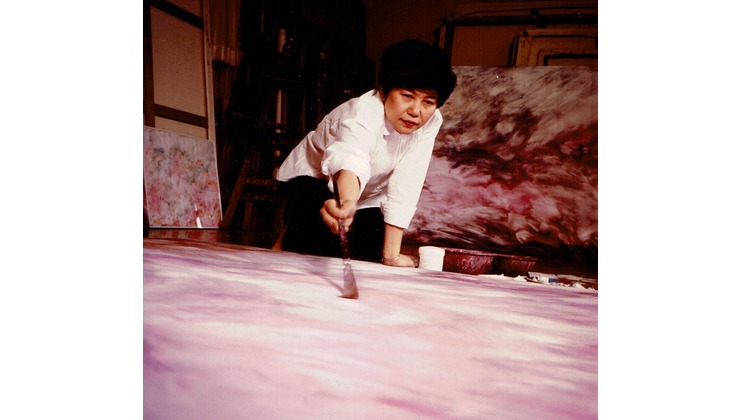 A person wearing a white shirt and dark pants is bending over, painting a large canvas on the floor with a brush. The canvas appears to have a pink and purple pattern. There are additional canvases and painting materials visible in the background.