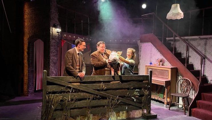 A theatrical stage set showing a scene with three actors. One man is holding a lamb, while the other two look on, smiling. The set includes a wooden pen, an old piano, a staircase, and a hanging lamp. The atmosphere is dimly lit with a misty haze.