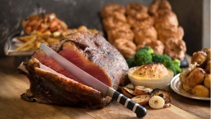 A platter of roast beef being sliced with a knife. The scene includes roasted vegetables, broccoli, and a small dish of a baked side. The background shows a tray of additional sides, including more vegetables and what appears to be a bread or pastry item.