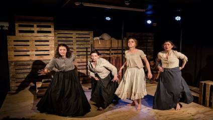 Four women are performing on stage, dressed in period clothing. The set is minimalistic, with wooden pallets and crates. The actresses seem engaged in an intense scene, with expressive body language and facial expressions in a dim-lit environment.