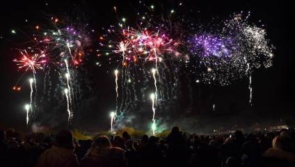 A large crowd watches an impressive fireworks display at night. Multiple colorful fireworks burst in the sky, illuminating the scene with vibrant red, green, purple, and white lights. The dark sky contrasts with the bright explosions, creating a festive atmosphere.