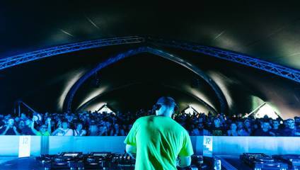 A DJ wearing a neon green shirt stands at a mixing console, performing for a large crowd under a large, dark, arched canopy. Blue lighting enhances the atmosphere, highlighting the audience's excited faces as they enjoy the music.