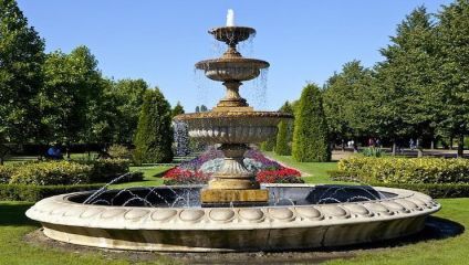 A large, ornate stone fountain with multiple tiers is seen in the middle of a lush park on a sunny day. Water cascades from the top tier into a circular basin surrounded by well-maintained greenery, trees, and flower beds with vibrant red blooms.