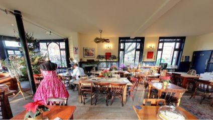 A cozy, eclectic café interior with mix-and-match wooden tables and chairs. A mannequin in a pink dress is on the left, plants and festive decorations are scattered throughout, and large windows let in natural light. A few patrons are seated.