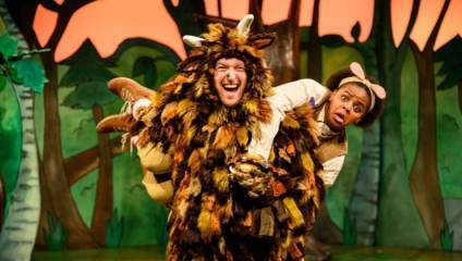 Two actors performing on stage in colorful costumes. One actor is dressed as a fantastical creature with brown, black, and white fur, lifting another actor dressed in an outfit with ears resembling a small animal. The background depicts a vibrant, cartoon-like forest.