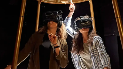 Two individuals are wearing virtual reality headsets, engaged in an immersive experience. The person on the left points forward while the person on the right reaches upward towards a hanging object. They are both indoors, under a structure with vertical supports.