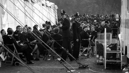 A black-and-white image shows numerous police officers in uniform seated on folding chairs and gathered outside a white tent. The scene appears to be a large, organized event, with several standing and conversing officers. The background shows more officers and spectators.