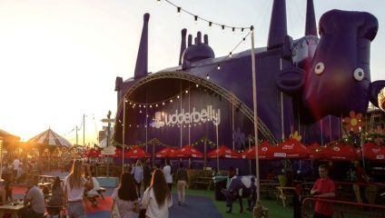 A large outdoor event venue featuring a giant purple inflatable structure with cartoonish features, labeled Udderbelly. People walk around and sit at tables under red umbrellas. The setting sun casts a warm glow over the lively scene.