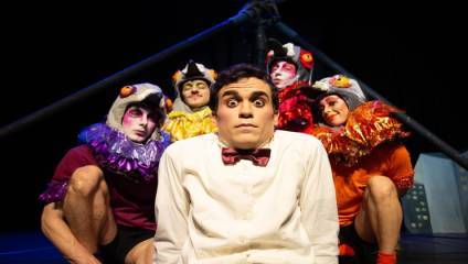A man in a white shirt and bow tie looks surprised and wide-eyed. Behind him, four performers in colorful bird costumes with exaggerated makeup crouch and pose against a dark background with a structure resembling a pole.