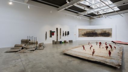 A modern art gallery with abstract installations. On the ground are various large, textured materials, including a piece with red shapes protruding. The walls display additional textured, unstructured art pieces. The space is well lit by ceiling lights.