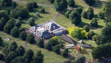 An aerial view of the Serpentine Gallery located in Kensington Gardens, London, surrounded by lush green trees and pathways. Adjacent is a uniquely designed pavilion with abstract geometric shapes and patterns, creating a striking contrast with the greenery.