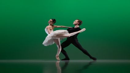 Two ballet dancers perform on stage against a green backdrop. The female dancer wears a white tutu and is on pointe, while the male dancer in black supports her with an extended arm. Both exhibit grace and poise in their positions.