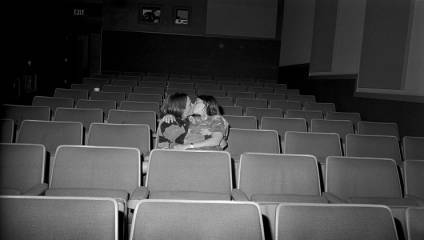 A black-and-white image of two people sitting in an otherwise empty theater. They are embracing and kissing, seated in the middle of the rows of empty seats. The setting is dimly lit with an exit sign visible to the left.