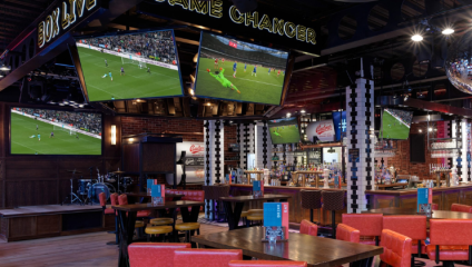 A sports bar with multiple large screens displaying live soccer matches. The bar features a mix of red and black seating, high-top tables, and a well-stocked bar. The atmosphere includes vibrant decor with sports-themed signs and a small stage with musical instruments.