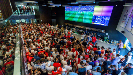 A large crowd of people, mostly dressed in red and white, are gathered in an indoor space, watching a soccer match on a large screen. Some are standing on a mezzanine level. The atmosphere appears lively and enthusiastic.