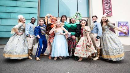 A group of people in colorful, elaborate costumes, likely for a theatrical production, stand happily in front of the Royal & Derngate theatre. They are posing with big smiles and enthusiastic expressions. The costumes range from regal and elegant to whimsical and playful.