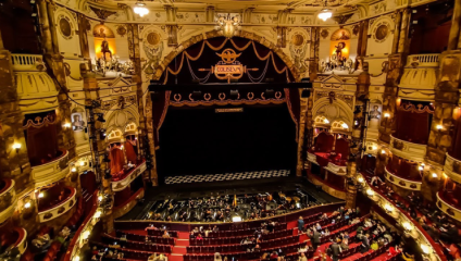 A grand theater interior with ornate decorations and elaborate lighting. The stage curtain reads Coliseum. Red seats fill the auditorium, some occupied by people. Below the stage, an orchestra pit with musicians is visible. The atmosphere is elegant and historic.