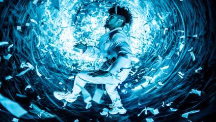 A person dressed in all white appears to be floating or jumping in a tunnel-like environment filled with swirling, illuminated blue lights and confetti. Their expression and posture suggest a dynamic, intense moment.
