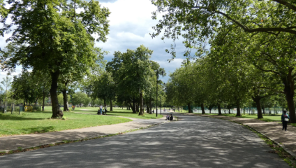 A serene park scene with a paved pathway winding through lush green trees and grassy areas. People are scattered along the path, sitting and strolling. The sky is partly cloudy, casting a mix of sun and shade across the landscape.