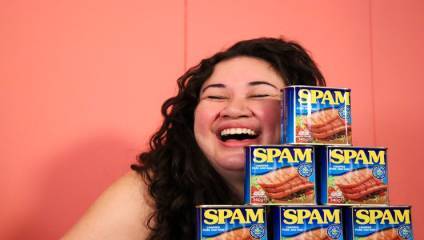 A woman with long dark hair laughs joyfully while posing next to a pyramid of six cans of Spam. The background is pink, and the cans are stacked in three levels, with two on the bottom, two in the middle, and one on top.