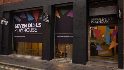 A building facade with large black signs displaying Seven Dials Playhouse in white text and colorful triangular shapes. The entrance has large windows, and a glass door on the right, both reflecting the street and adjacent structures.
