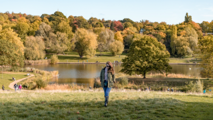 A woman stands on a grassy hill in a park, wearing a green jacket and blue jeans. Behind her is a calm lake surrounded by autumn-colored trees. People are walking along the paths, enjoying the scenic view under a clear sky.