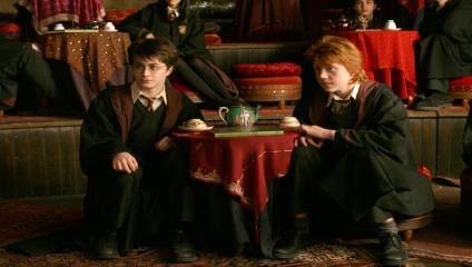Two young people wearing black robes with maroon linings sit beside a small table with a red cloth, a teapot, and teacups. Behind them, other robed individuals sit at similar tables adorned with patterned cloths and red cushions. The setting has a cozy, dimly lit ambiance.