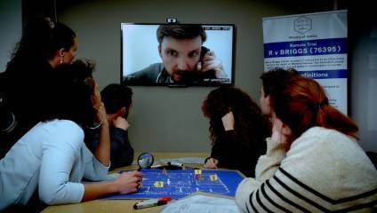 A group of people are gathered around a table, watching a man on a video call displayed on a wall-mounted monitor. The table holds papers and a map with markers. A banner nearby reads Ministry of Justice, mentioning Remote Trial and providing case details.