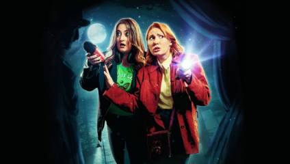 Two women, one wearing a green shirt and black jacket and the other in a red jacket, stand in a dark, misty environment, illuminated by the moonlight. They both hold flashlights, looking startled as they search the surrounding area.