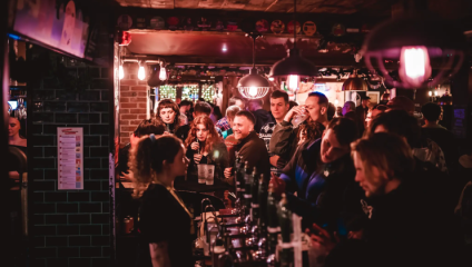 A dimly lit, crowded bar filled with people socializing and enjoying drinks. Patrons are gathered at the bar counter, chatting and drinking, with colorful lights and decorations enhancing the lively atmosphere. The background has brick walls and various wall hangings.