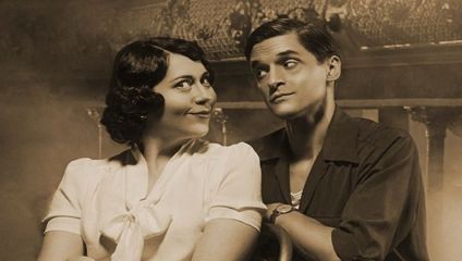 Sepia-toned image of a woman and a man with vintage-style clothing and hair. The woman, on the left, smiles and looks at the man. The man, on the right, smirks and looks back at her with a playful expression. The background features ornate decorations.