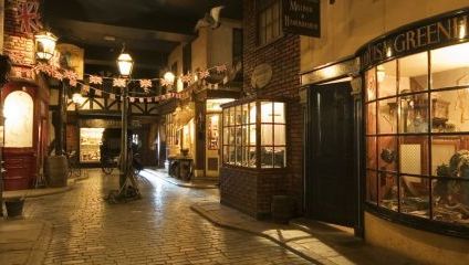 A dimly lit, old-fashioned street scene with cobblestone pavements and vintage shopfronts. The shops display items in their windows, and bunting with Union Jack flags hangs above. Antique street lamps illuminate the scene, giving it a nostalgic ambiance.