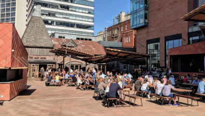 A group of people are sitting at outdoor tables on a sunny plaza surrounded by modern and rustic buildings. Many are conversing and enjoying food or drinks under a clear blue sky. The area has a relaxed and lively atmosphere.
.