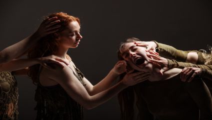 A group of people participates in a dramatic scene. A red-haired woman on the left places her hand on another woman's head, who is screaming with her eyes closed as multiple hands touch her face. The background is dark, emphasizing the intense emotional expression.