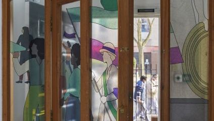 A wooden door with glass panels features colorful stained glass artwork. The designs depict abstract figures, including a woman with a sword and shield, and other people in various poses. Sunlight illuminates the artwork, casting vibrant hues and creating a welcoming atmosphere.