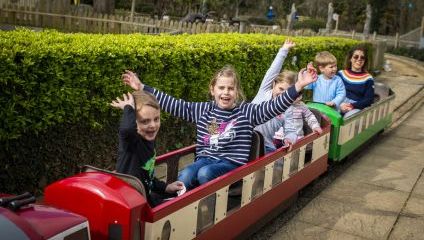 A group of happy children and an adult are riding a small outdoor train. The children are smiling and waving their hands in the air. The train is red and green, and they are surrounded by lush greenery with trees and bushes. Everyone looks excited and cheerful.