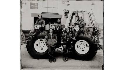 A group of five individuals dressed in similar protective suits and helmets stand and sit around a large vehicle with oversized tires. They pose confidently in front of a building with stairs, suggesting a serious and professional context. The photo is in black and white.