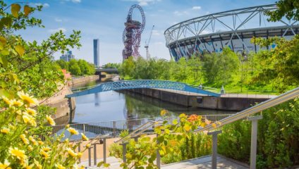 A scenic view of Queen Elizabeth Olympic Park in London, featuring the ArcelorMittal Orbit sculpture, the London Stadium, a tree-lined river with a blue pedestrian bridge, and vibrant greenery and flowers in the foreground. Bright, sunny day with blue skies.