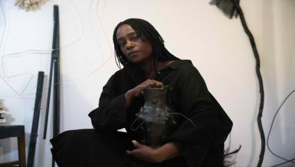 A person with long braided hair, wearing a dark outfit, holding a sculptured dark vase, sits in an interior space with various abstract items and artworks around. They have a thoughtful expression and are slightly leaning forward. The lighting is soft and natural.