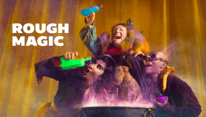Three people in vibrant costumes enthusiastically mix colorful potions into a steaming cauldron, with smoke rising from it. The background is a warm yellow-orange, adding to the magical atmosphere. The text ROUGH MAGIC appears on the left side of the image.