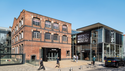 A brick building labeled Science and Industry Museum with a modern glass extension, featuring a large sign that reads Cafe. People walk near the entrance, and parked cars are seen on the right. The sky is clear and blue.