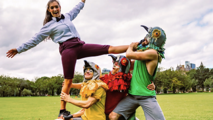 A woman in a blue long-sleeve shirt and purple pants balances on the shoulders of a person in a yellow shirt wearing a bird-like mask. Two others, also wearing bird masks, in green and red shirts, support her. They are outdoors on a grassy field with trees in the background.