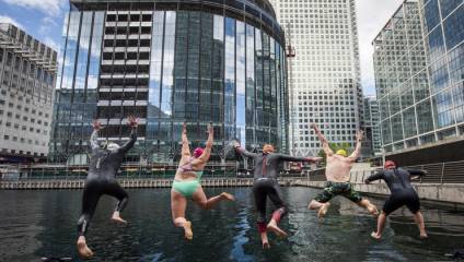 A group of five swimmers, wearing wetsuits and swim caps, jump into the water together in an urban area surrounded by modern high-rise buildings and glass structures. One swimmer wears a green swimsuit instead of a wetsuit. The sky is partly cloudy.