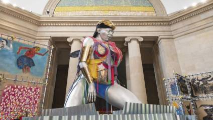 A large, brightly adorned sculpture of a woman, featuring metallic and colorful elements, stands in the center of a grand hall with classical architecture. Surrounding the sculpture are various pieces of colorful, abstract artwork.