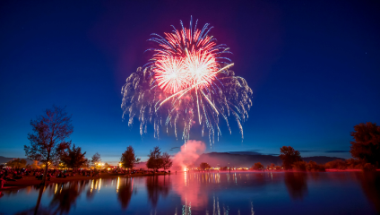 A vivid fireworks display lights up the night sky with bursts of red and white over a calm lake. The reflections of the fireworks and nearby lights create a serene and festive atmosphere, with silhouettes of trees and people along the water's edge.