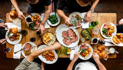 A group of six people sits around a wooden table enjoying a meal. The table is filled with plates of roast beef, potatoes, vegetables, and various drinks. Hands are reaching for food and passing dishes, indicating a lively dining scene.