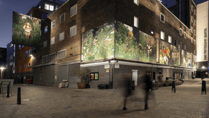 A nighttime photo of a brick building adorned with large, illuminated photographs that depict various garden scenes. Two blurred figures walk along the cobblestone street in the foreground. The sign Soho Photography Quarter is visible on the side of the building.